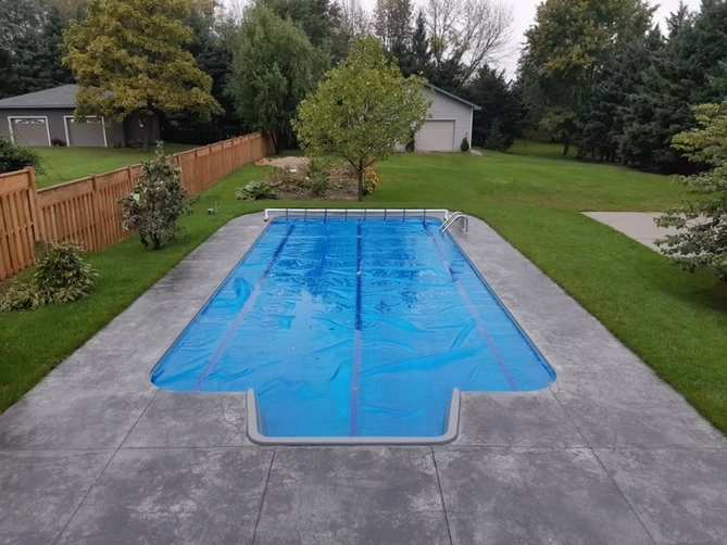 Rectangular in-ground swimming pool with a safety cover, stamped concrete deck, and spacious landscaped backyard.