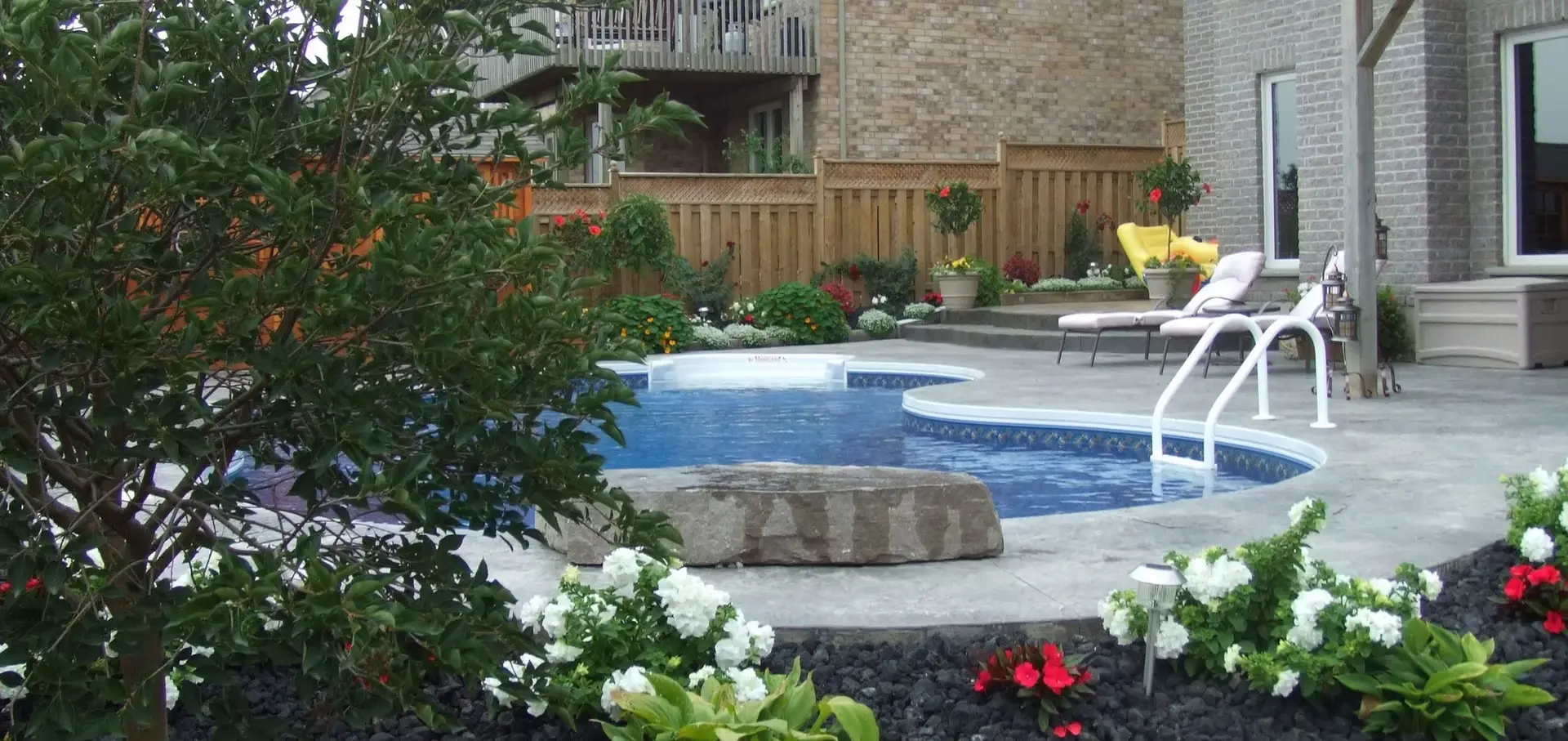 Custom freeform inground pool with a textured concrete deck, large natural stone accent rock, and lush poolside flower beds