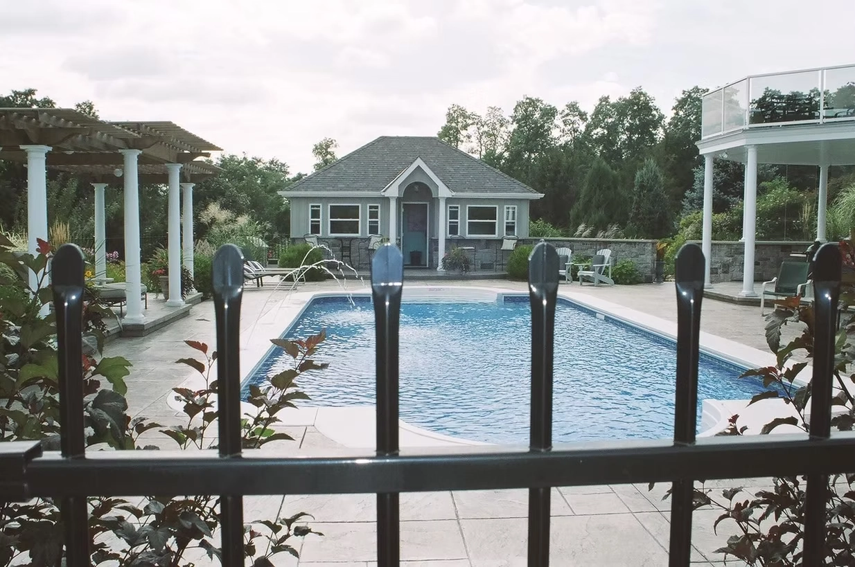 Luxury backyard swimming pool with decorative water features, pergola, pool house, and safety fence.