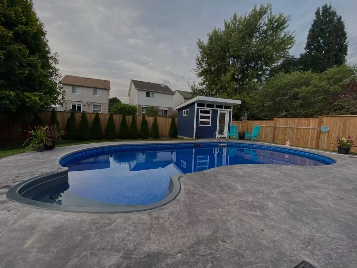 Freeform in-ground swimming pool with stamped concrete patio, pool shed, and fenced residential backyard.